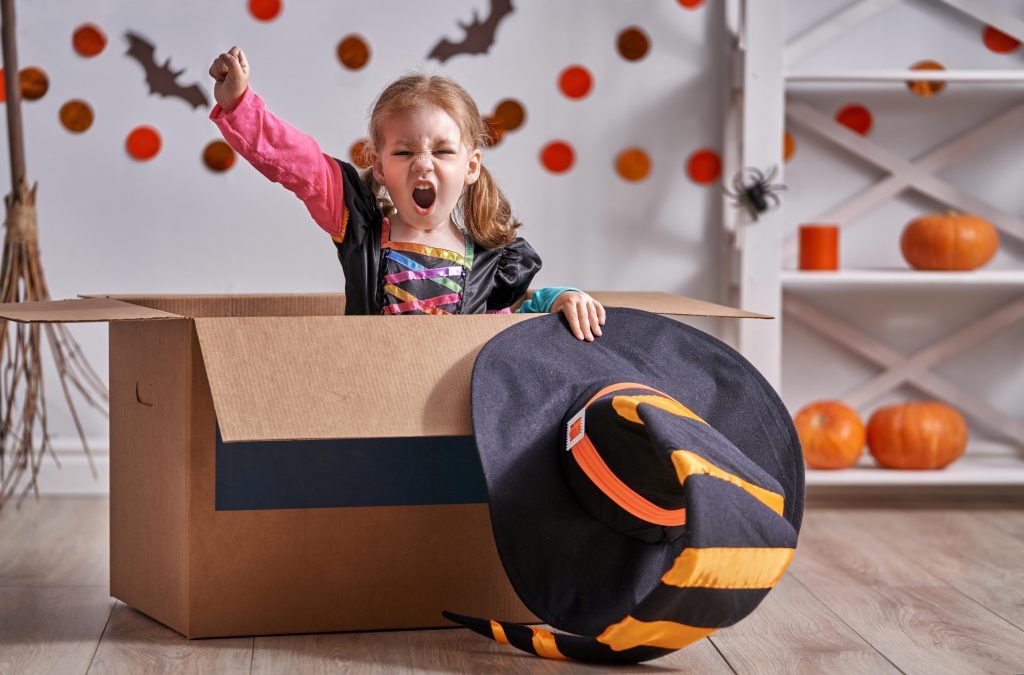 little girl sits in cardboard box cheering while wearing halloween witch costume with halloween decorations in background