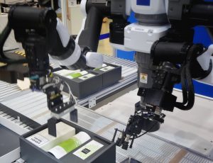 two collaborative robot arms place hair care products into black boxes on conveyor line